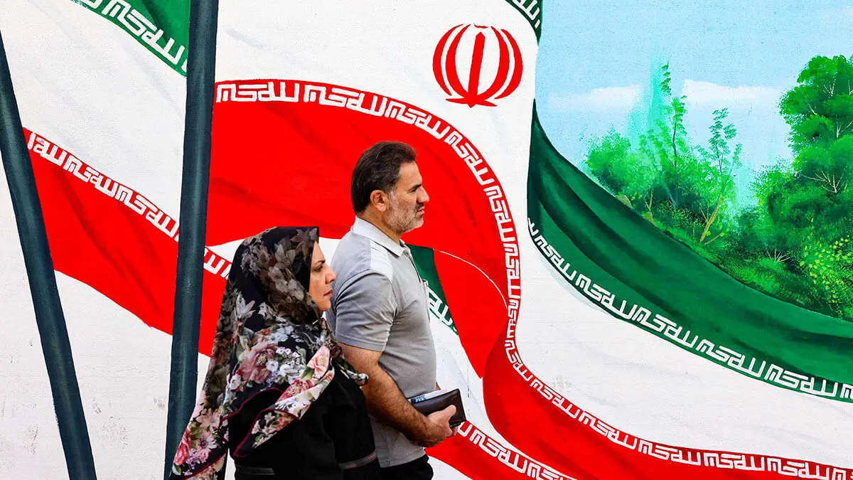 <p>Un hombre y una mujer pasan junto a un mural con banderas nacionales iraníes en una calle de Teherán - AFP/ATTA KENARE </p>