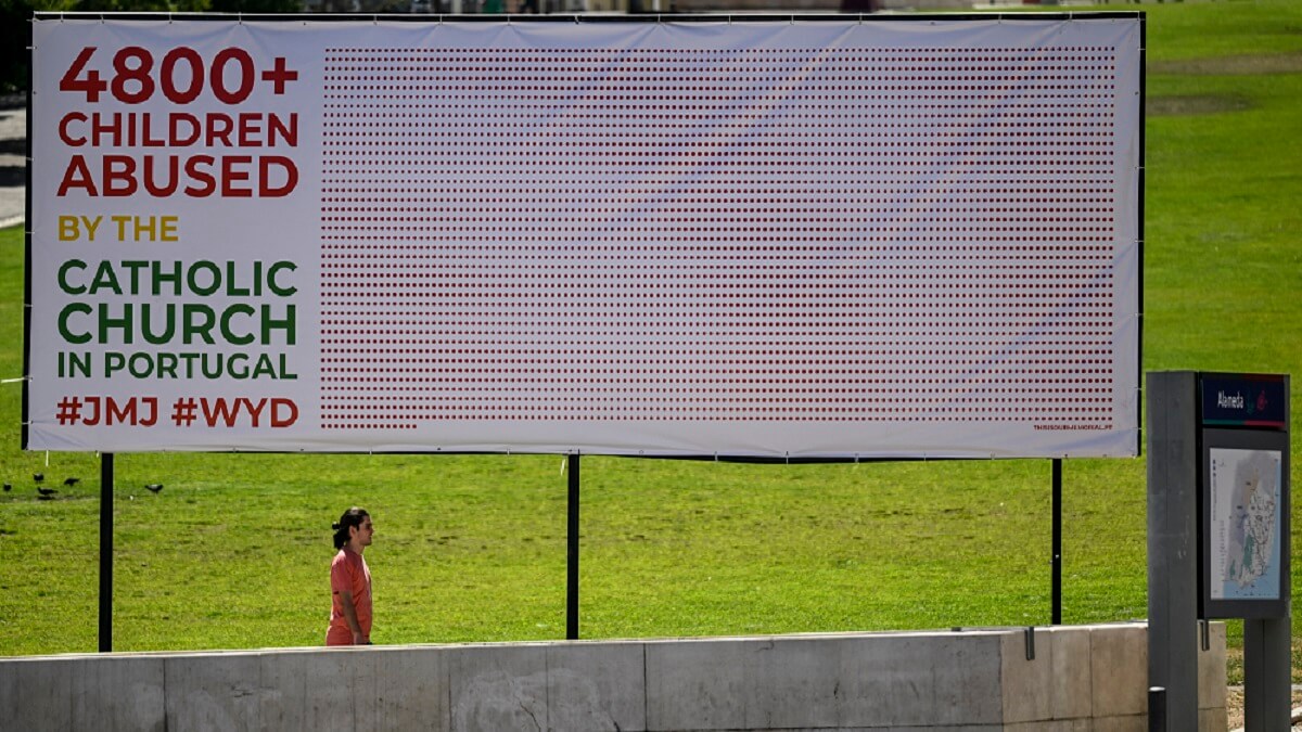 PHOTO/AFP/PATRICIA DE MELO MOREIRA - Un hombre pasa junto a una valla publicitaria que denuncia los abusos sexuales cometidos por miembros de la Iglesia católica portuguesa en la Alameda de Lisboa el 2 de agosto de 2023, durante las celebraciones de la Jornada Mundial de la Juventud (JMJ)